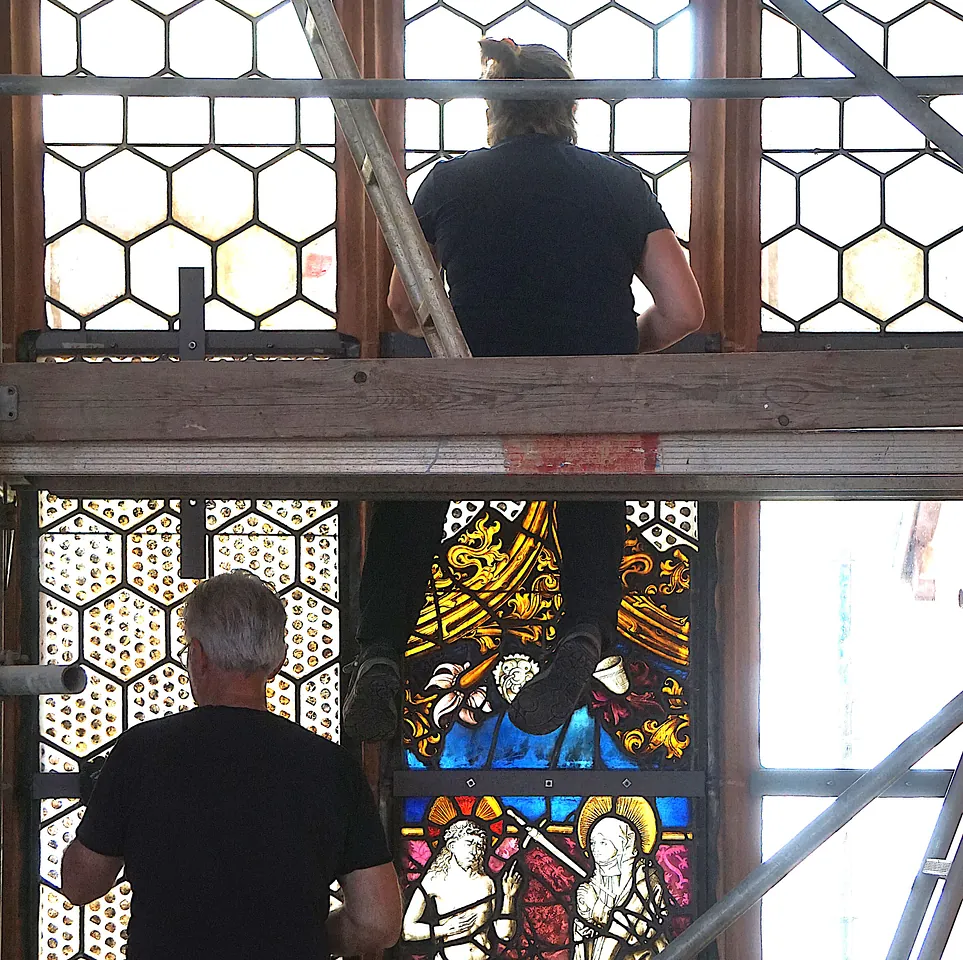Zwei Menschen auf einem Baugerüst bauen die restaurierten Fenster wieder ein.